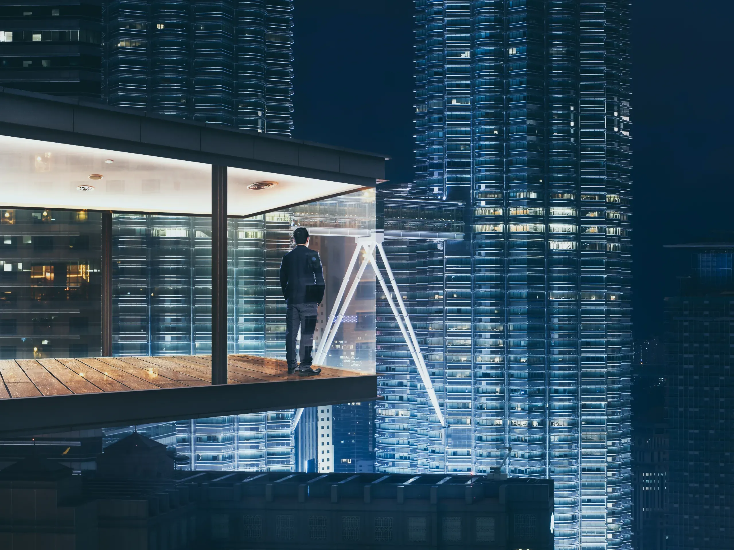 Businessman in a modern office at night looking out over an illuminated city skyline through floor-to-ceiling windows. 