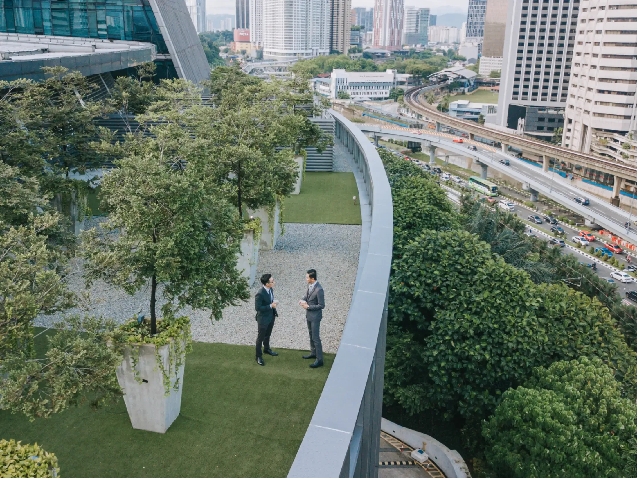 Two business colleagues talking in a rooftop garden outside an office building, with modern architecture and city views in the background. 